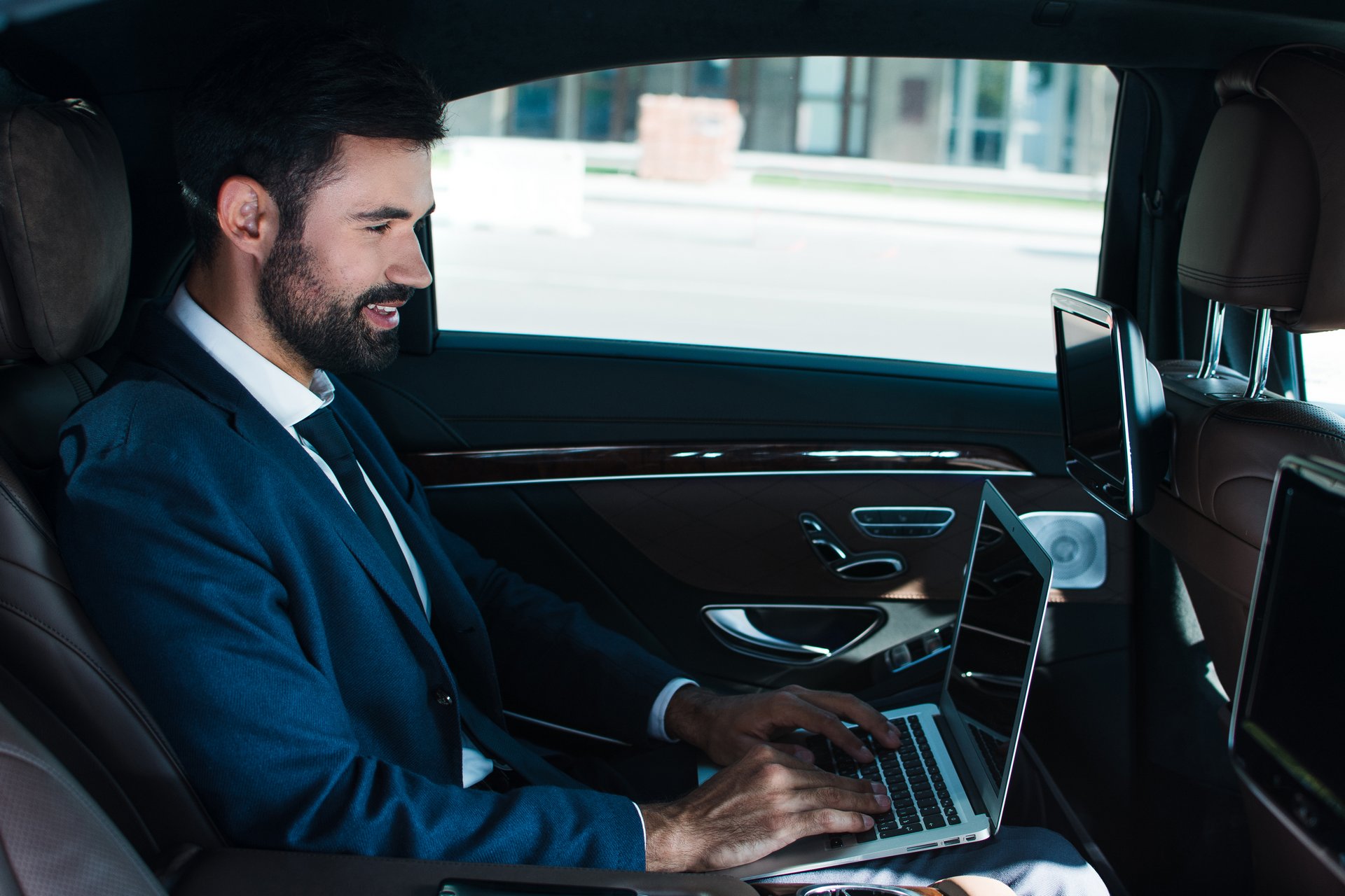 On the way to successful meeting. Side view of handsome young man using laptop with smile while sitting in car